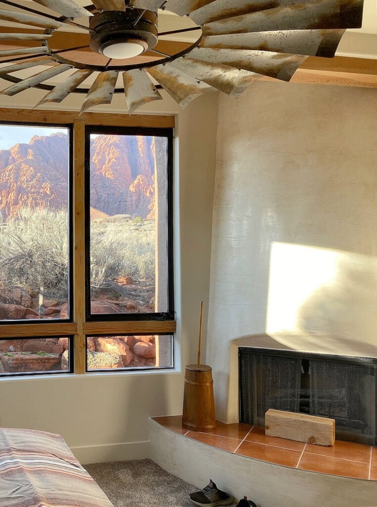 corner drywall fireplace with limewash beige walls, wood window, tile hearth, desert view out the window, rustic industrial style fan in bedroom