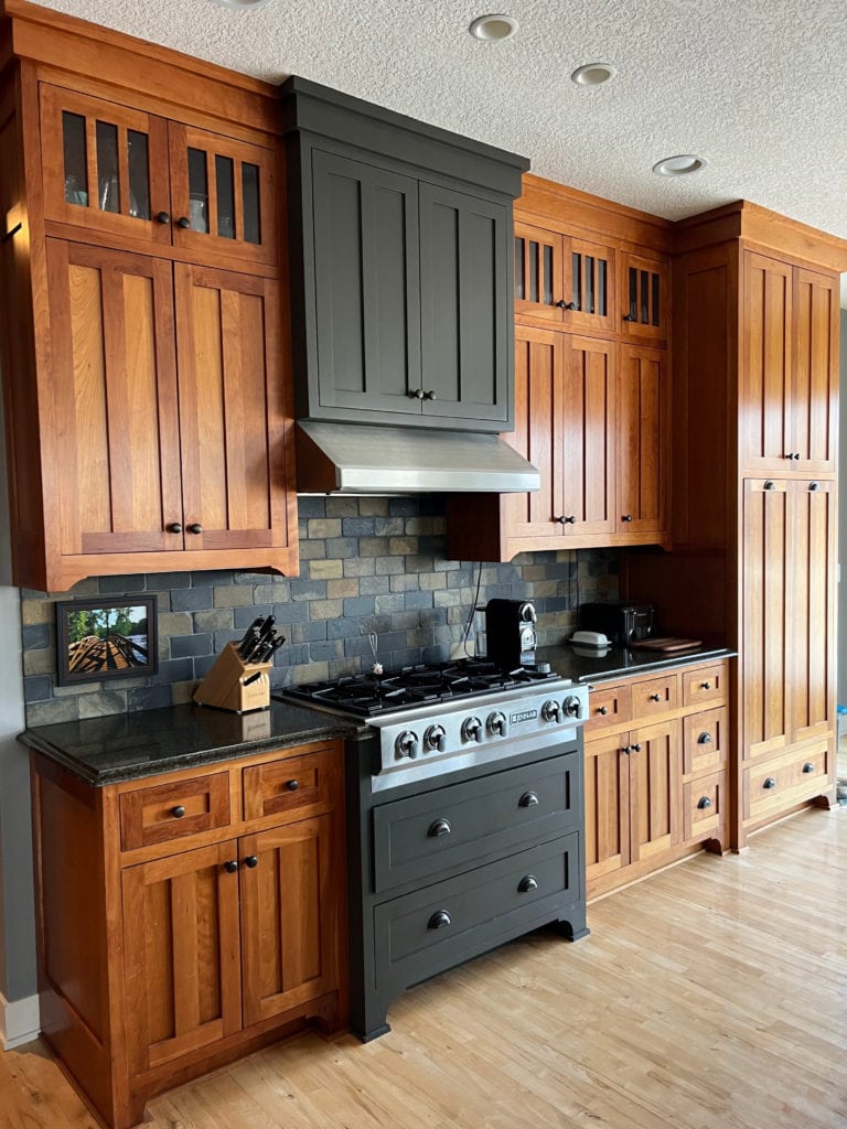 kitchen with slate tile backsplash, black granite countertop, dark gray green painted hood fan and cherry wood cabinets. Kylie M Interiors