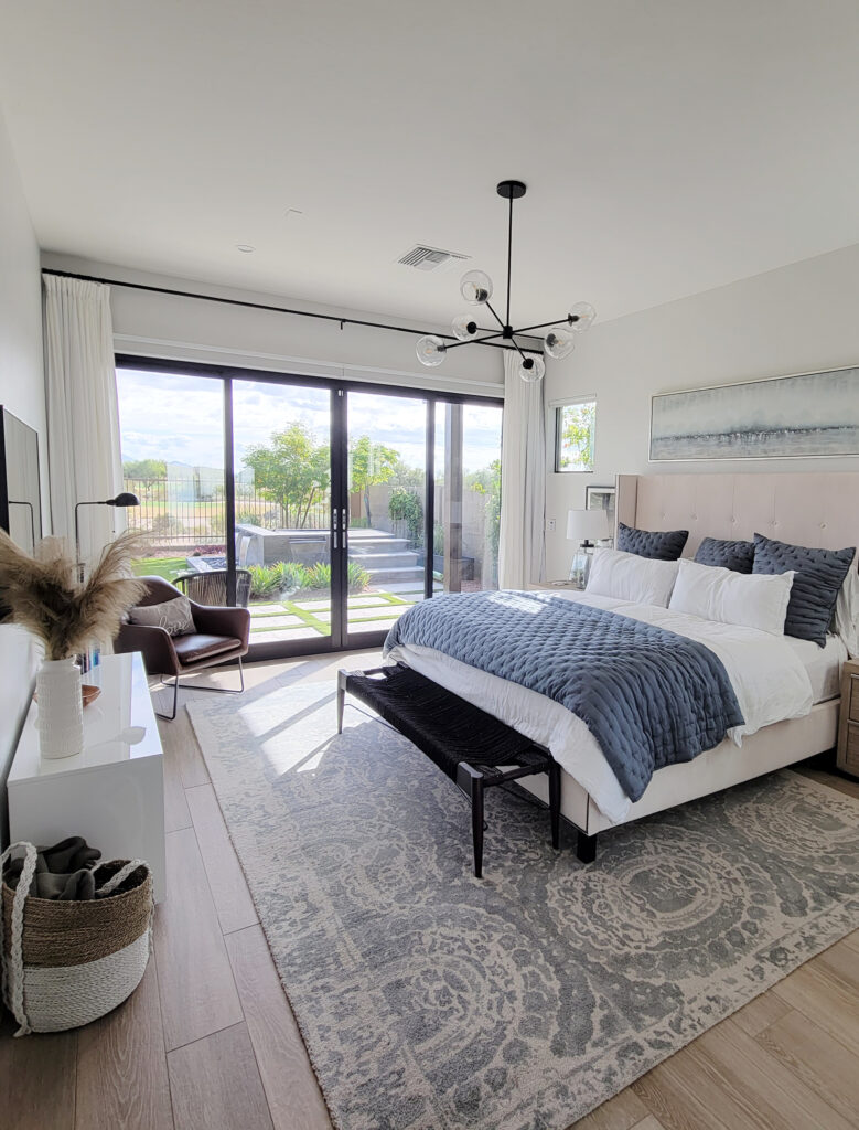 Primary bedroom walls Dunn Edwards Faded Gray similar to Benjamin Moore Silver satin, area rug on wood, blue accents, sputnik chandelier.