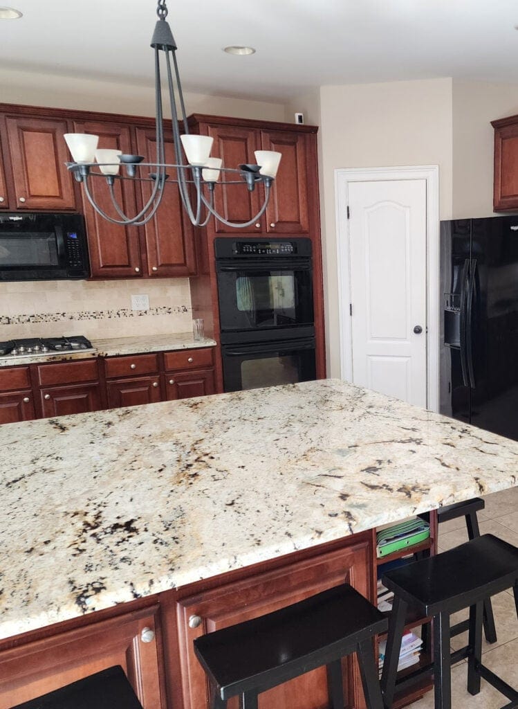 Cherry red wood stained kitchen cabinets, granite countertop with beige, gold, taupe, cream, beige tile floor, travertine tile backsplash, black appliances