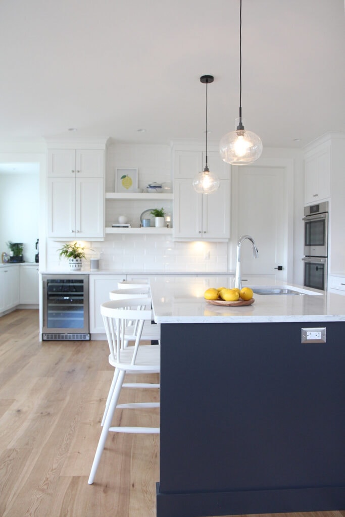 Kitchen with oak wood flooring, Goodfellow, door to pantry, island Sherwin Williams Cyberspace, navy blue, white cabinets in Pure White, floating shelves, wine fridge, white quartz