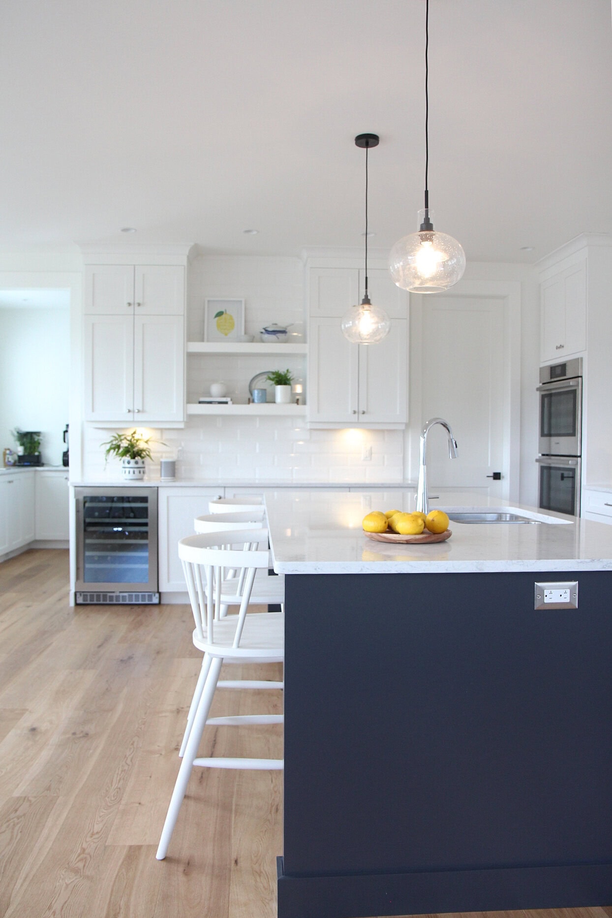 Kitchen with oak wood flooring, Goodfellow, door to pantry, island Sherwin Williams Cyberspace, navy blue, white cabinets in Pure White, floating shelves, wine fridge, white quartz