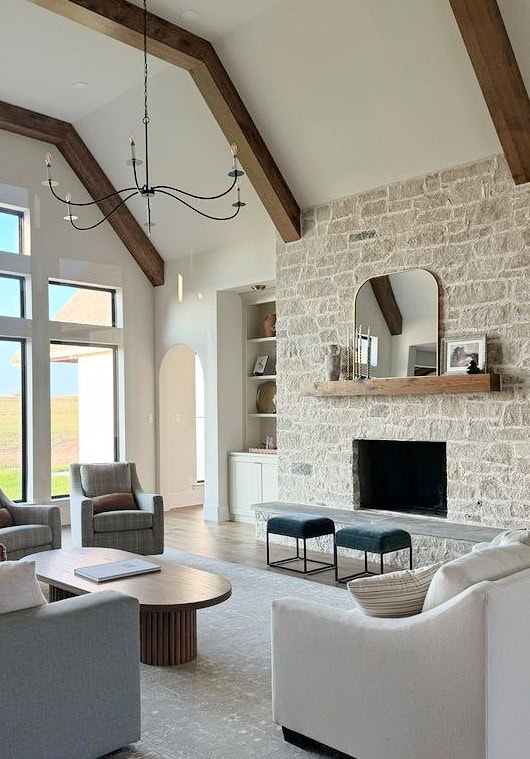 Off-white Sherwin Williams paint color on walls, vaulted living room ceilings, wood beams, warm cream stone fireplace, wood mantel, sofa, chairs, arched doorway