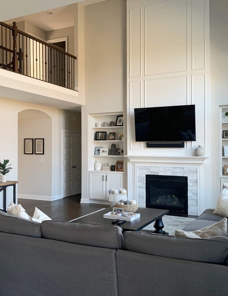 Benjamin Moore Super White trim, moldings on vaulted high ceiling fireplace with marble, Sherwin Williams Agreeable Gray walls, gray sectional, wood floor, open railing catwalk. Built-in, archway