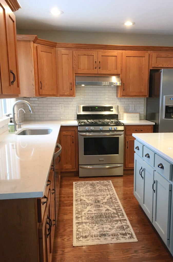 Kitchen with orange-red, possible red-oak kitchen cabinets, wood flooring, blue-gray painted island in Sherwin Williams Comfort Gray, white quartz countertop, white subway tile, update ideas