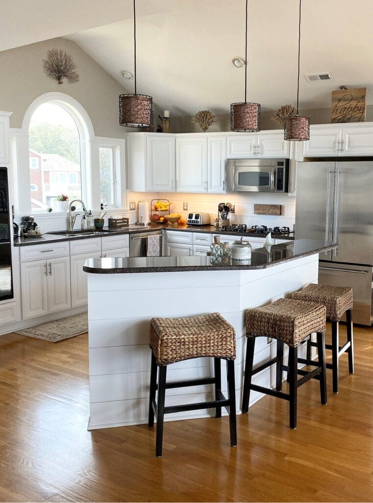 Kitchen with painted warm white cabinets, similar to Benjamin MOore White Dove, dark granite countertop, golden oak wood floor, shiplap back of island and backsplash, rattan stools
