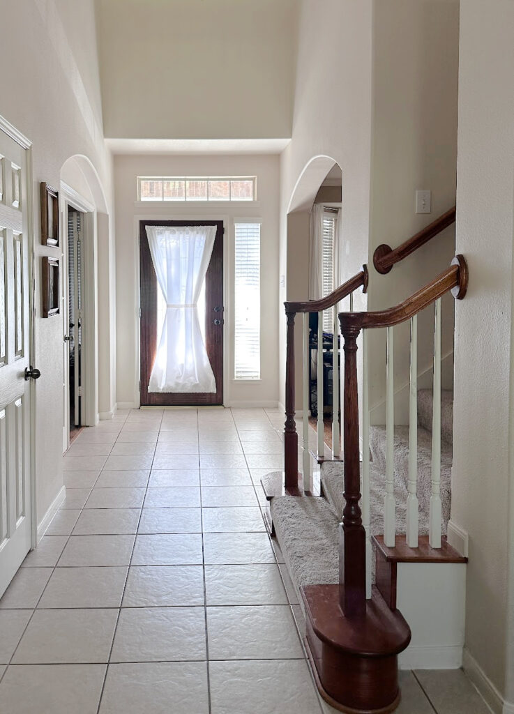 Sherwin Williams Moderate White in hallway with 12x12, 1990s tile flooring and cherry red wood stair railing. High ceiling. Update ideas for 2000s staircase.