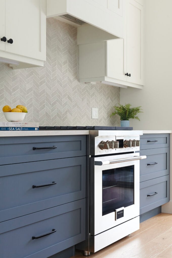 fulgor appliances, Benjamin Moore Brittania Blue lower cabinets, island, White dove walls and uppers, chevron tile backsplash, Omnia quartz countertop, black hardware, white oak