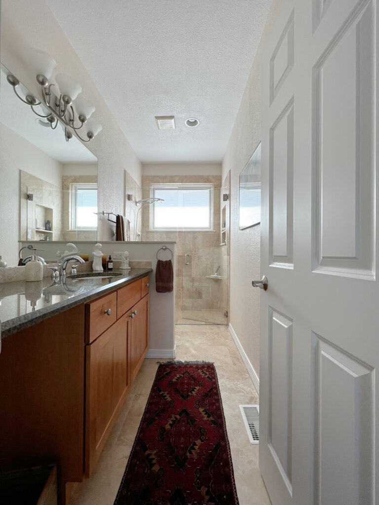 Bathroom with travertine tile floor, shower surround, wall paint color Sherwin Williams Divine White, dark granite countertop, wood vanity