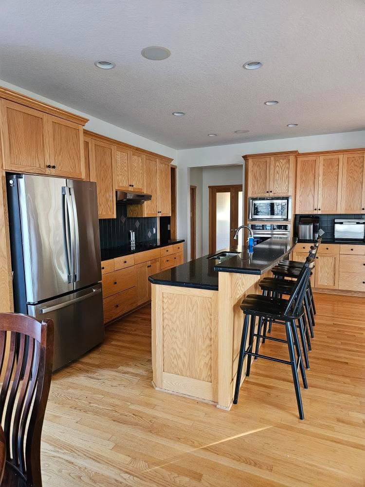 Oak wood kitchen cabinets, island with raised bar, black granite countertop, stainless steel appliances, golden oak floor