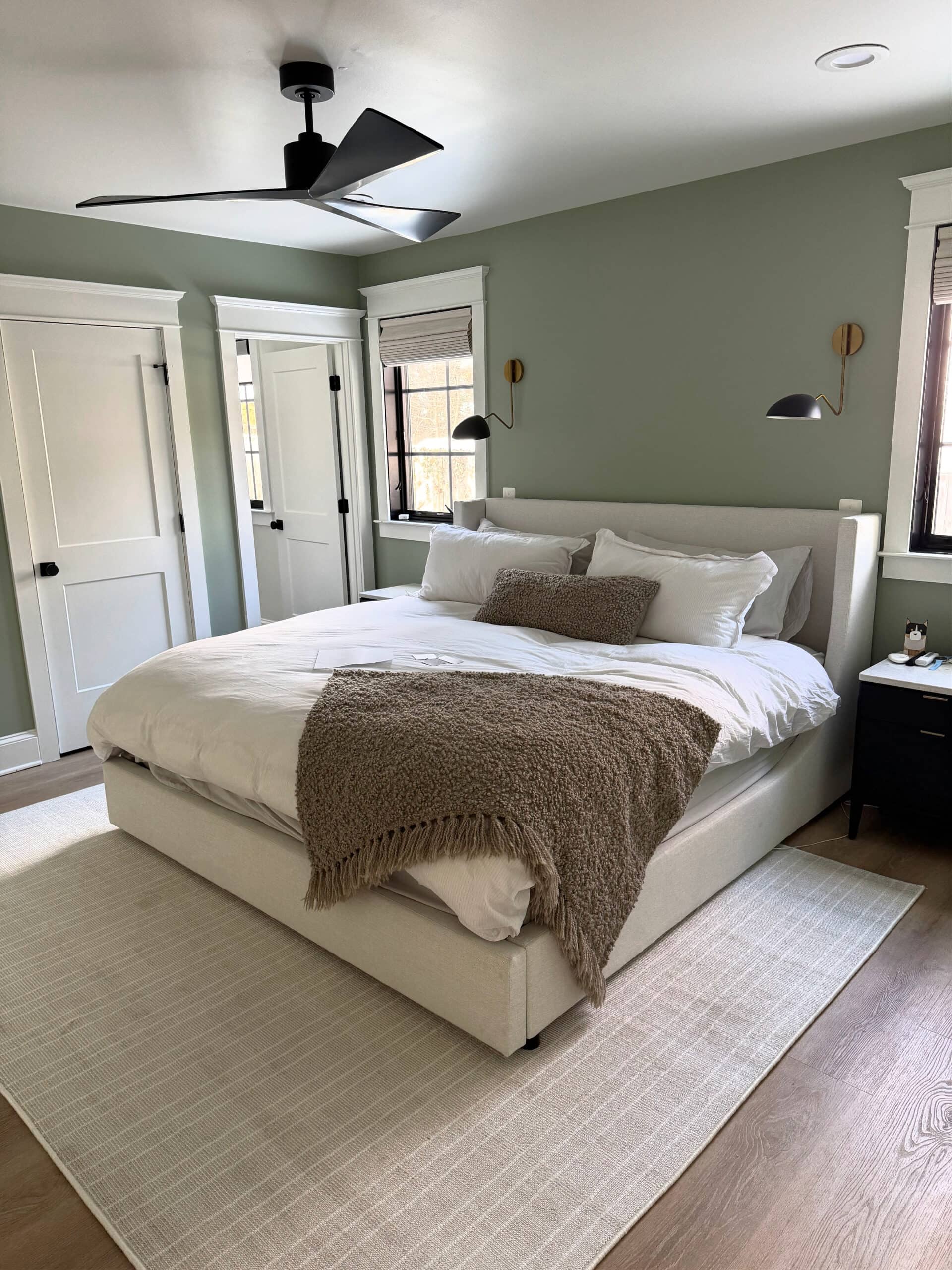 Behr Ultra Pure White trim, ceilings, doors, primary bedroom painted the best green paint color, north facing, Sherwin Williams Cascade Green, linen upholstered bed frame and headboard, laminate floor, area rug, wall mount light sconce