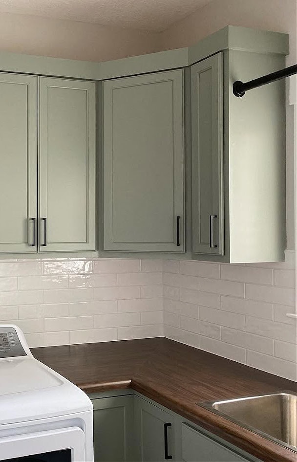 Green painted laundry room wood cabinets similar to Sherwin Williams Evergreen Fog, butcher block countertop, subway tile backsplash, white washer, black hardware