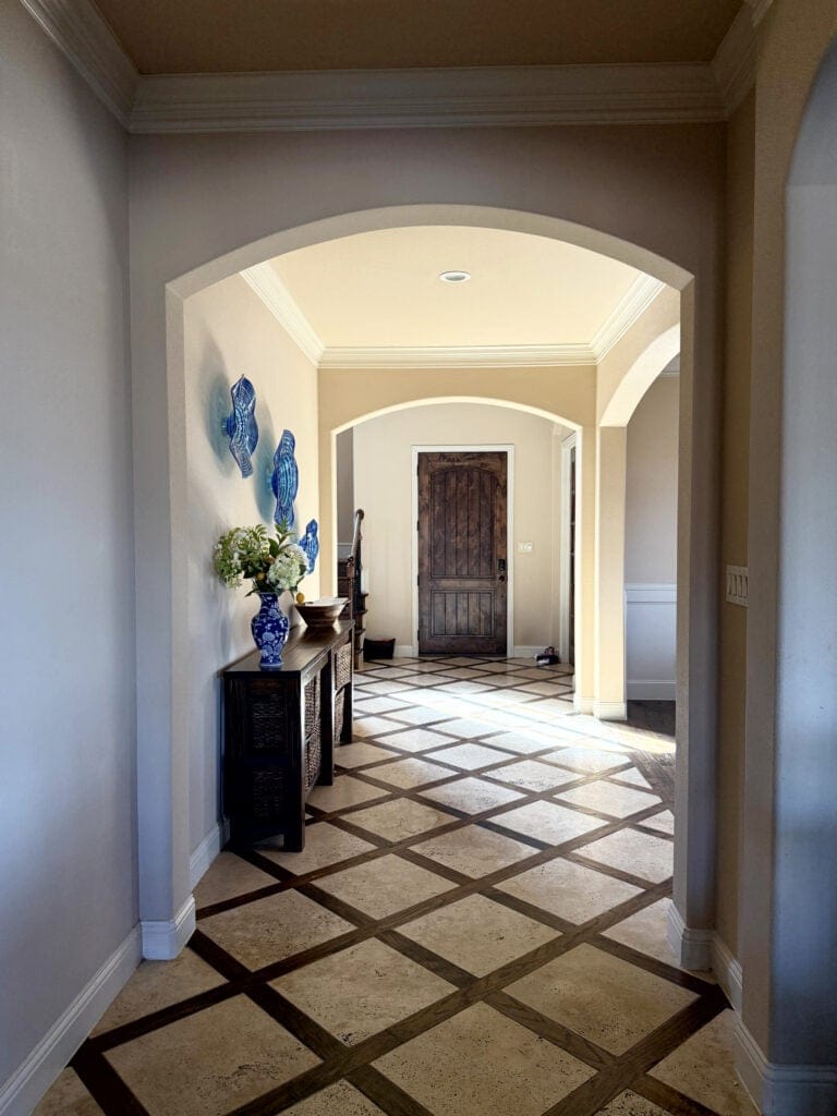 Hallway with trellis or checkerboard pattern travertine tile floor, dark wood border, Similar to Sherwin Williams Kilim Beige walls, off-white warm cream trim, arched doorways