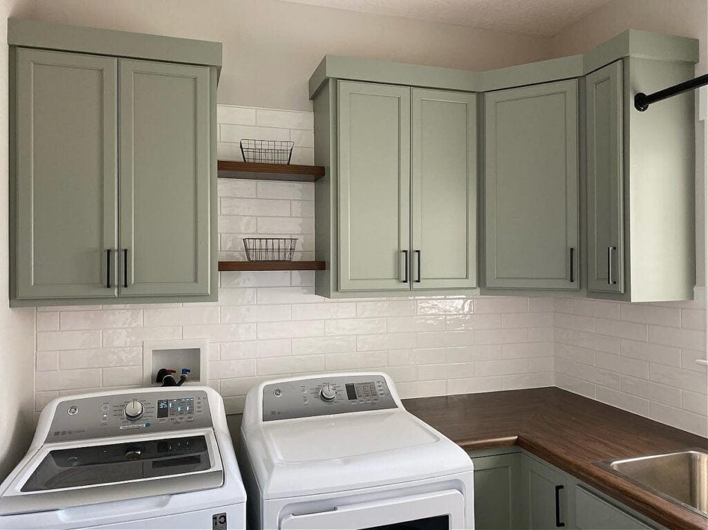 Similar to Sherwin Williams Evergreen Fog green laundry room painted cabinets, butcher block wood countertop, white subway tile, floating shelves, white washer and dryer