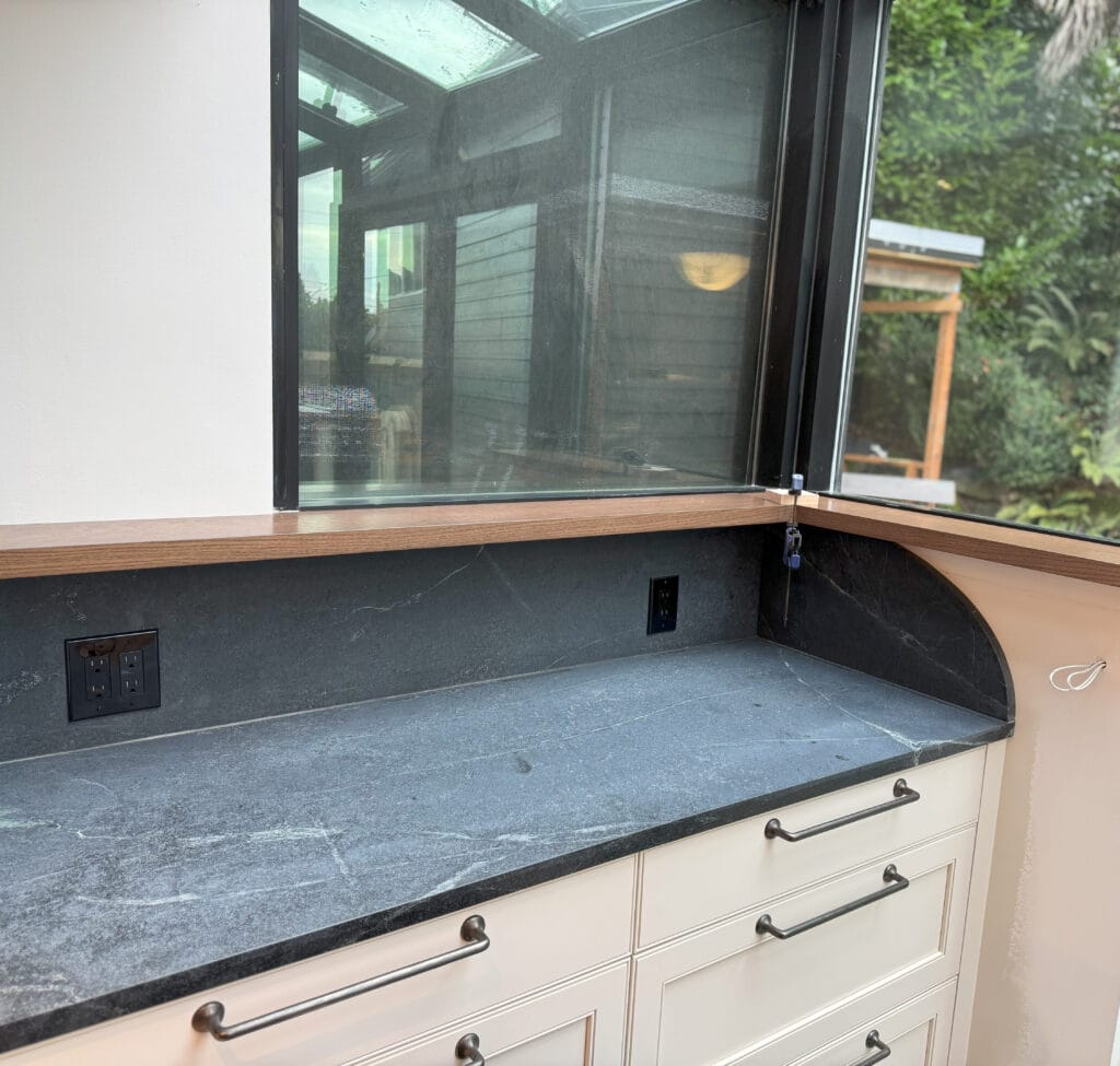 Kitchen with Benjamin Moore Wind's Breath off white painted cabinets, pewter hardware handles, black and green soapstone countertop, wood window ledge