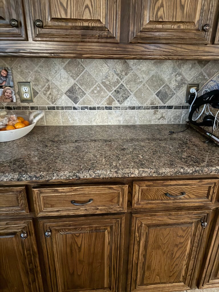 Dark oak wood cabinets, baltic brown look granite countertop, beige brown travertine tile backsplash in outdated dark kitchen