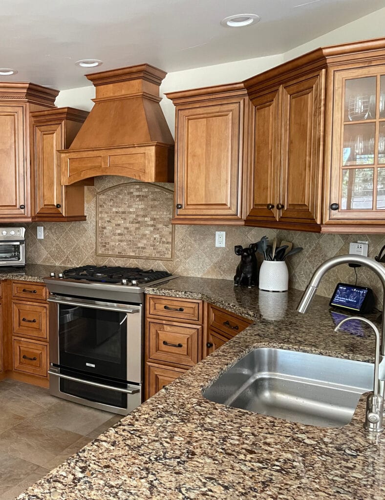 Stained orange wood maybe maple cabinets, beige tile floor, granite countertop early 2000s travertine look backsplash tile, stainless steel appliances. (1)