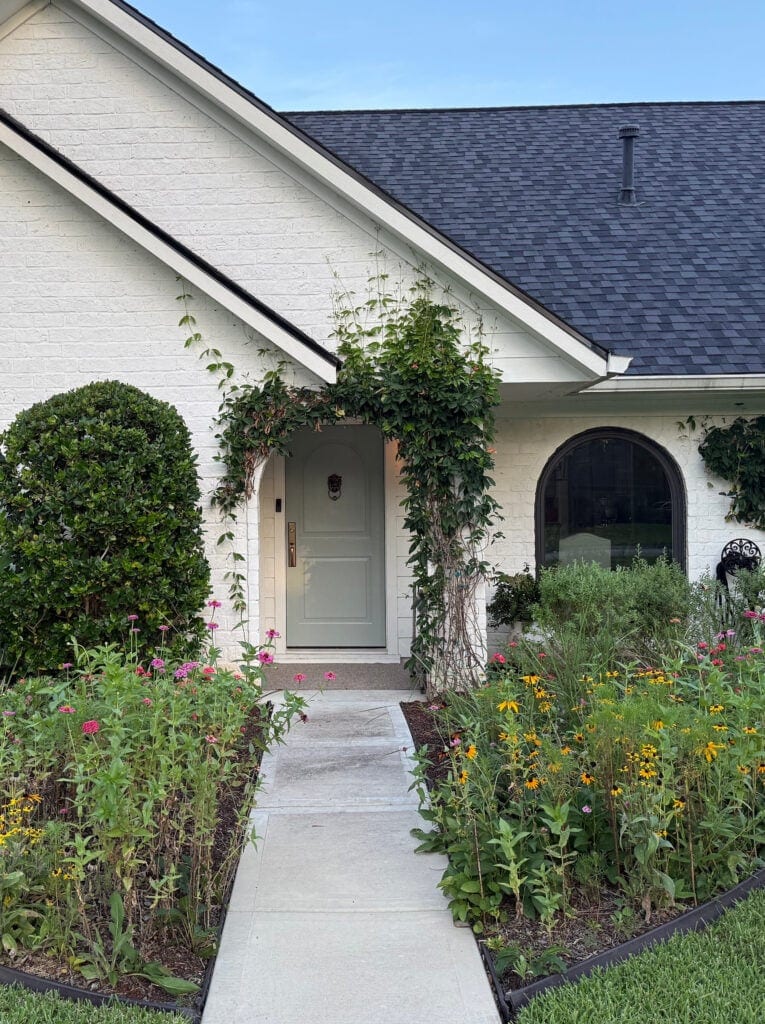 Sherwin Williams Oyster Bay green gray front door paint color with cottage style home painted brick in off-white Romabio finish with dark black charcoal roof and front garden